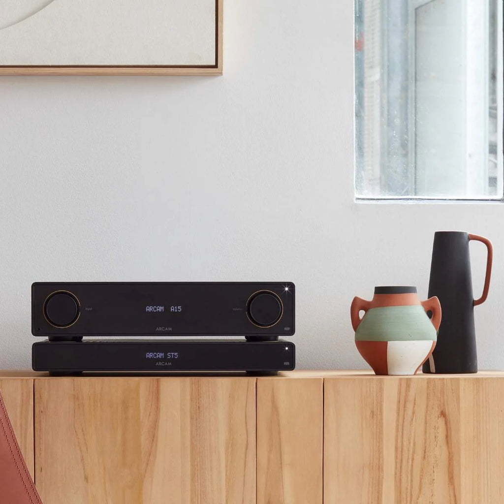 Arcam A15+ stereo amplifier in a stylish black finish, shown in a lifestyle setting with audio equipment and home decor.
