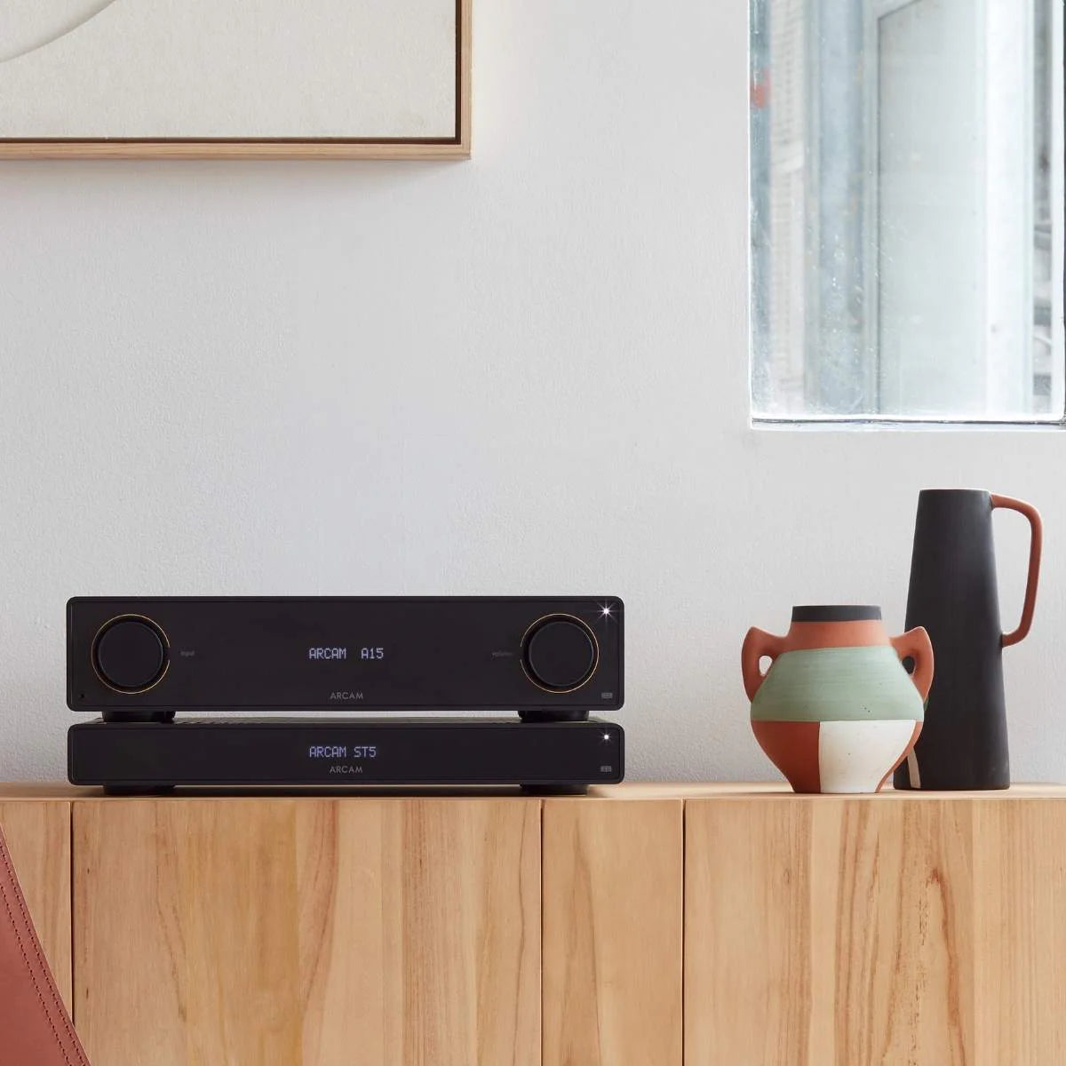 Arcam A15+ stereo amplifier in a stylish black finish, shown in a lifestyle setting with audio equipment and home decor.