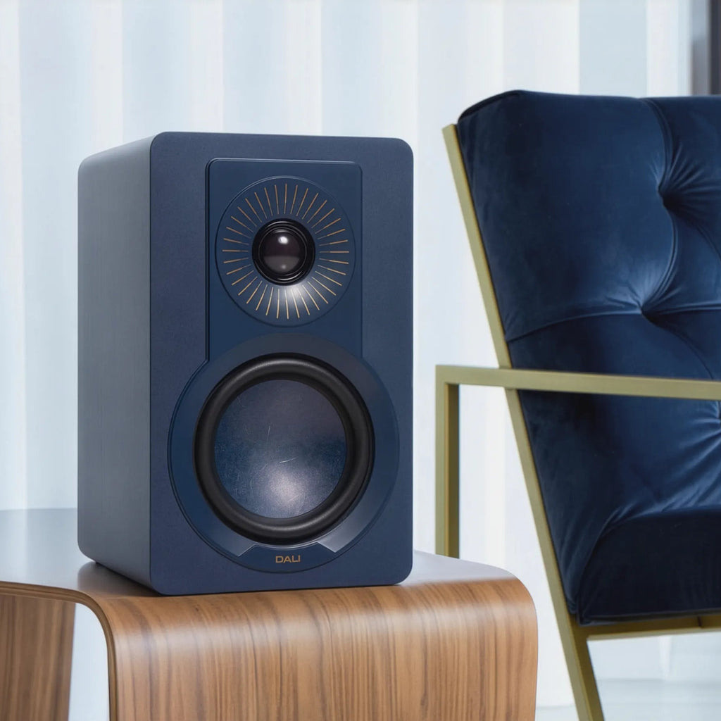 DALI Kupid blue passive bookshelf speakers on a table, showcasing compact design and speaker drivers in a modern home setting.