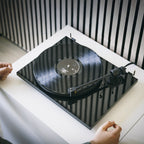Pro-Ject E1.2 BT High Gloss Black turntable playing a record on a white console, hands at the deck