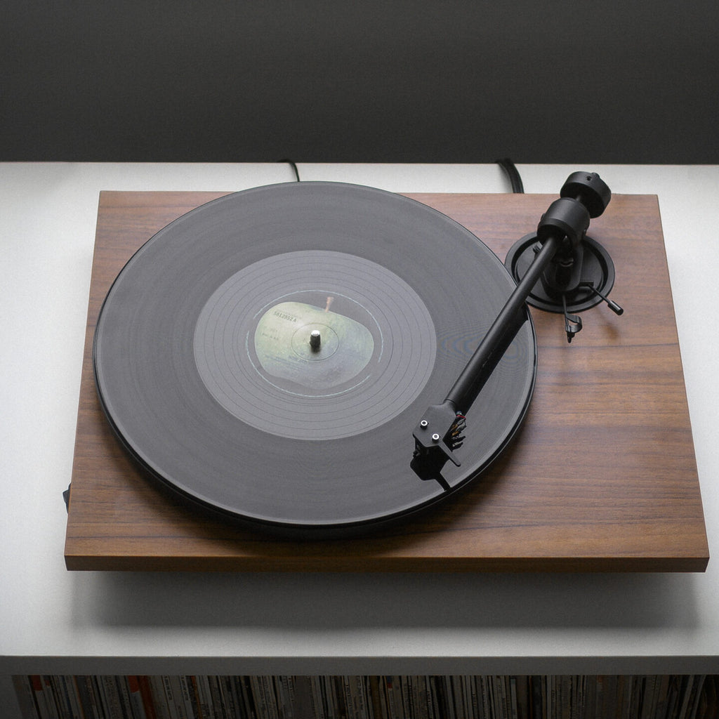 Pro-Ject E1.2 BT Walnut turntable playing a record on a console above a record collection